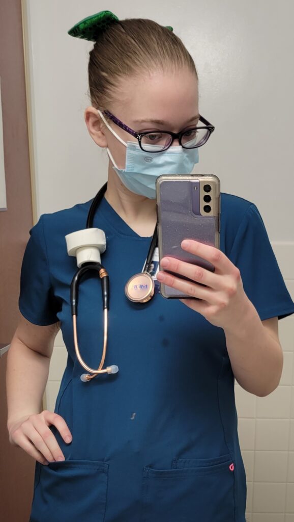 Nurse in Carribean Blue scrub top with stethoscope around neck. Green bow in hair taking selfie.