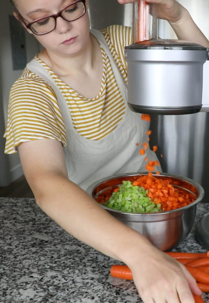 Stand Mixer with Dicer Attatchment with metal bowl, carrots, and celery. Girl in Yellow Striped Shirt and Apron.