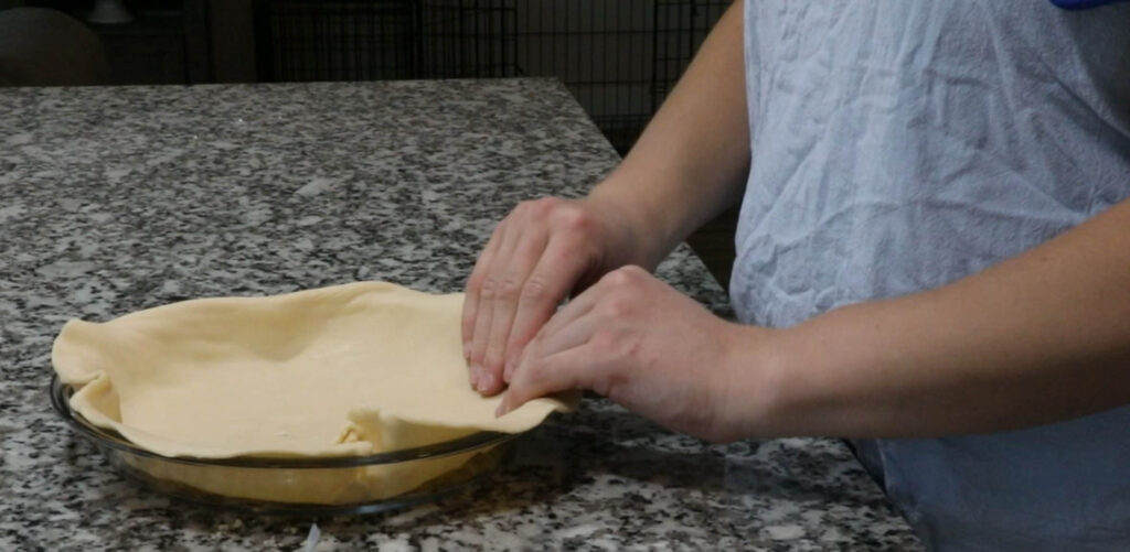 Pressing Pie dough into pie plate. Woman wearing blue apron in kitchen.