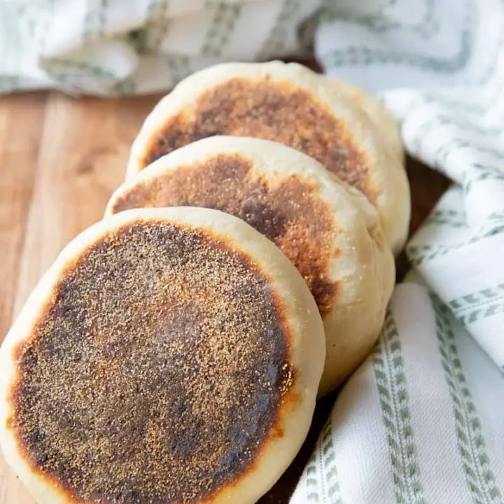 English muffins stacked on each other. Sitting on wooden cutting board with green fabric.